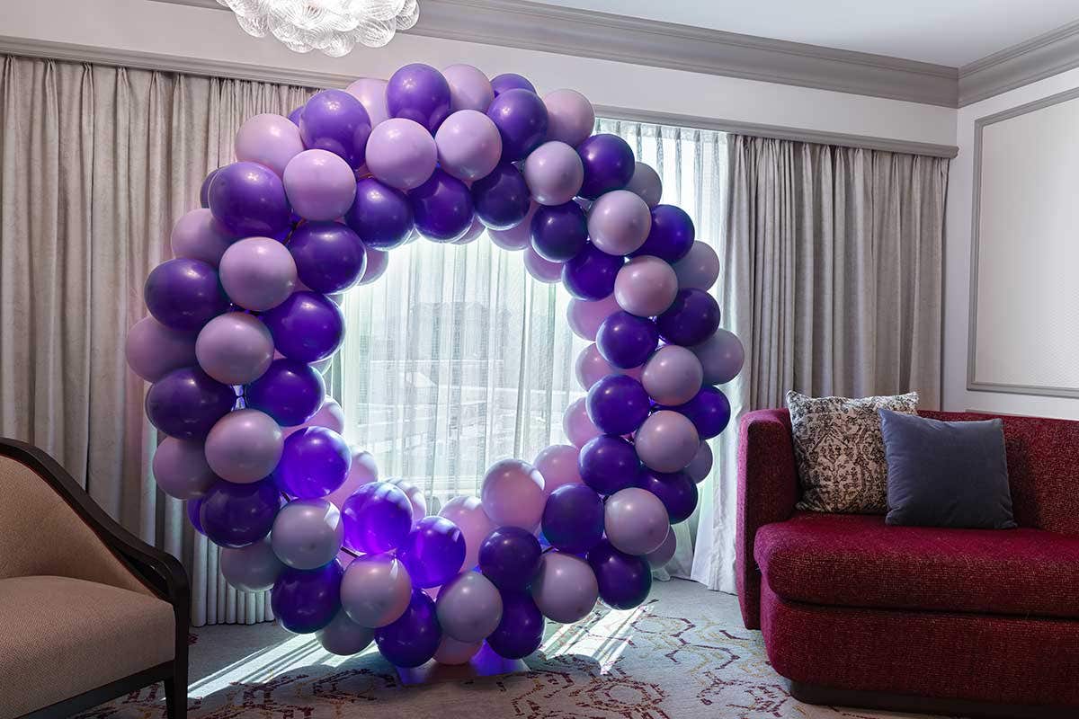 Large circular balloon arrangement in shades of purple and lavender displayed indoors, set in front of a window with sheer curtains, with a red upholstered chair and neutral-toned seating nearby.