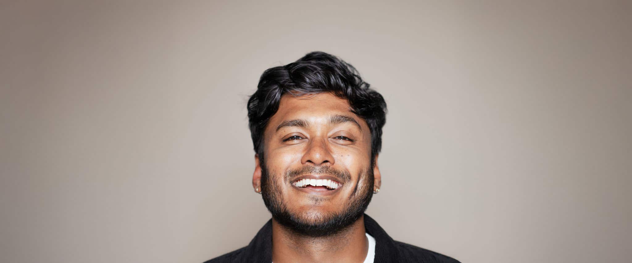 Usama Siddiquee in a white shirt and black jacket smiling at the camera in front of a beige background. 