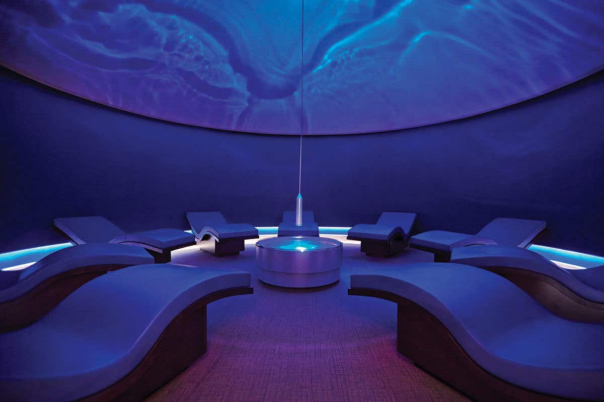9 relaxed day beds sit in a circle around a center table. The room is bathed in blue light as a light hangs from the middle of the room projecting water reflections.