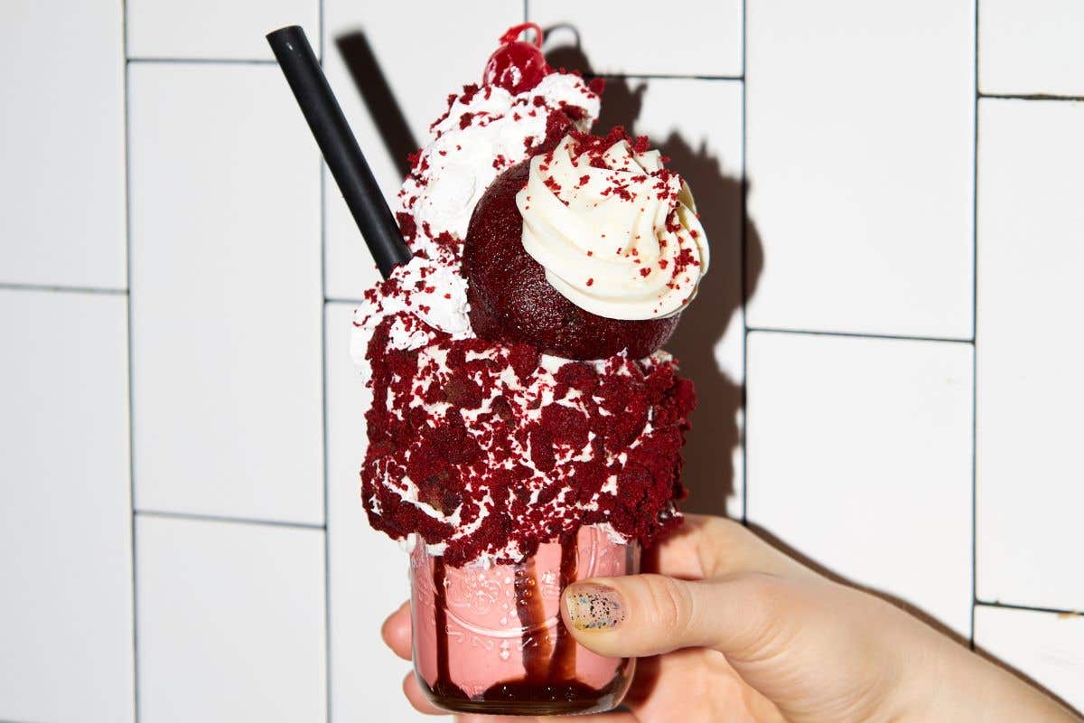 Hand holding a red velvet–themed shake with whipped cream.