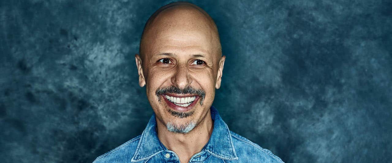 Maz Jobrani in denim shirt smiling and looking away from the camera standing in front of smokey blue background.  