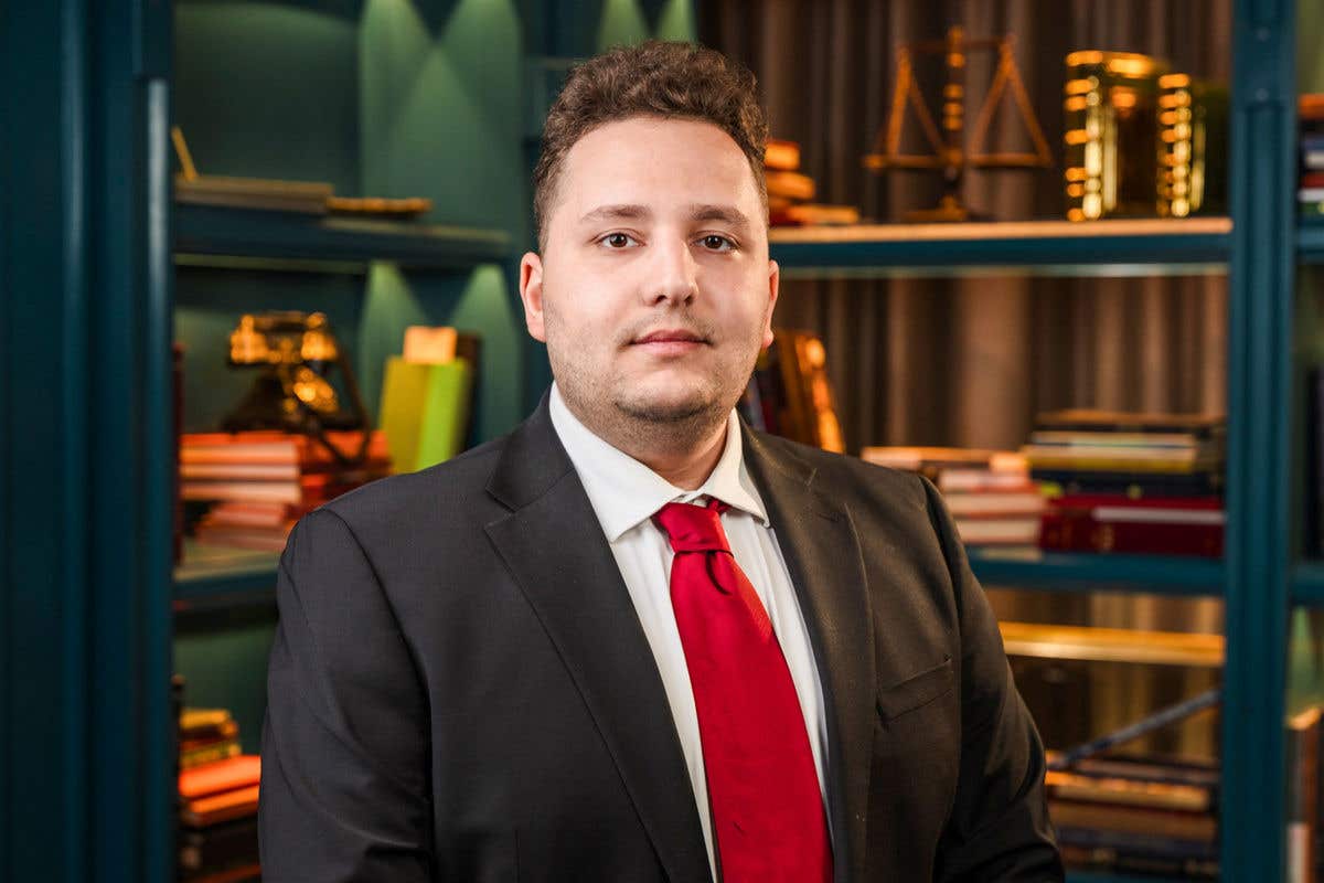A professional headshot of a man with wavy dark hair, wearing a black suit and a bright red tie. The background features teal bookshelves with books and vintage objects.