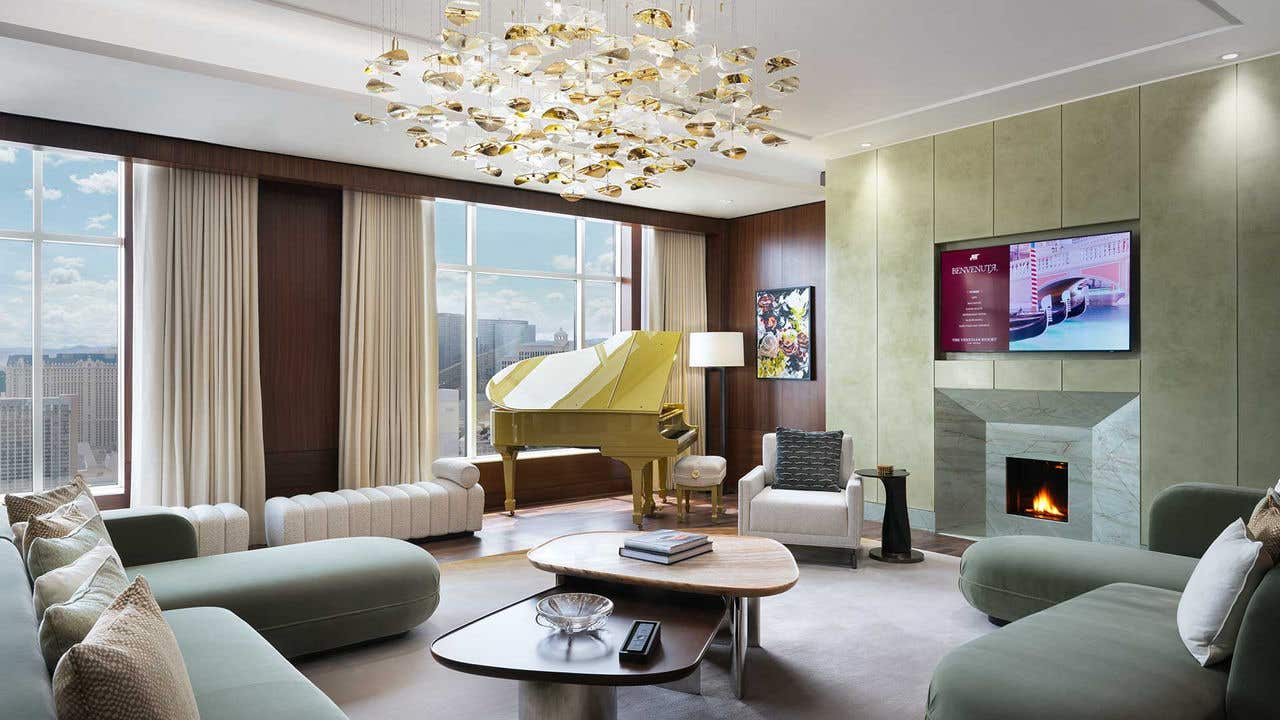 A large hotel living room with a chandelier, couches, armchairs, a gold baby grand piano, fireplace, TV, and a view of the Las Vegas Strip.