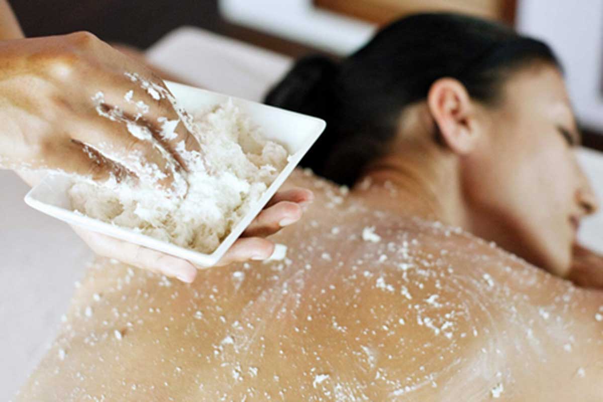 A bowl with white body scrub is held on the left by two hands as it is administered to a woman’s back who is lying face-down. 