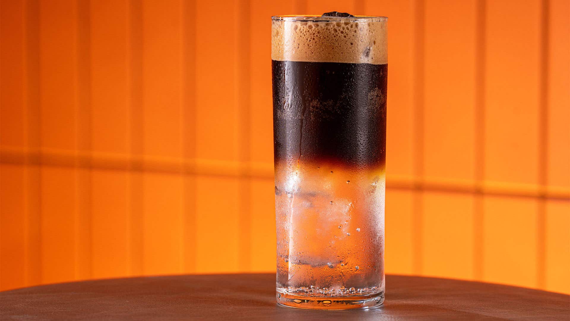 A cocktail in a tall glass, with brown liquor on top and clear liquor on the bottom on a table with an orange background.