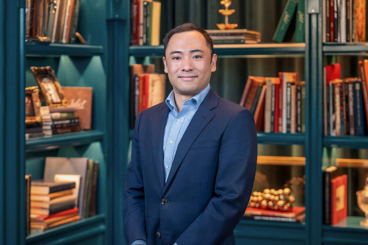 A professional headshot of a man with short dark hair, wearing a navy-blue suit jacket over a light blue button-down shirt. He is posed against a teal bookshelf background filled with books and vintage decor.