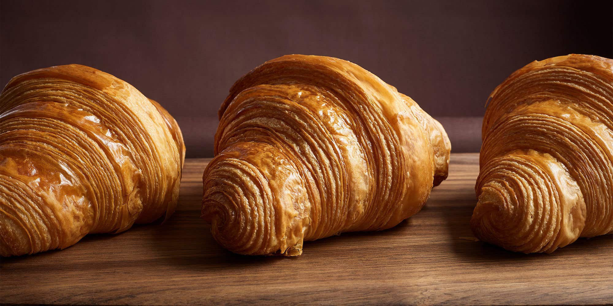 Three golden, flaky croissants arranged in a row on a dark wooden board.