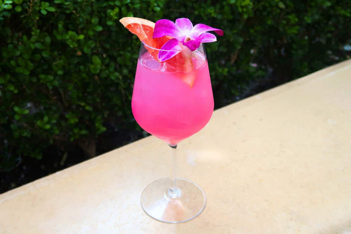 Bright pink cocktail served in a stemmed glass, garnished with a slice of grapefruit and a purple orchid flower, sitting on a light-colored table with green foliage in the background.