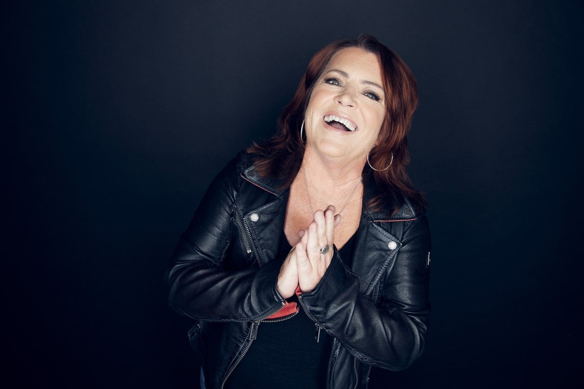 A woman with red hair in a black leather jacket smiles brightly with her hands clasped together against a dark background.