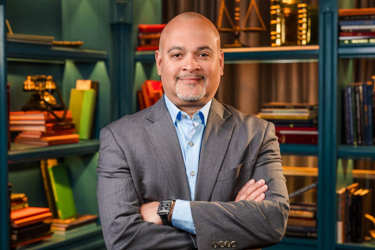 A professional headshot of a man with a goatee, smiling with his arms crossed. He is wearing a grey suit jacket over a light blue shirt and a watch. The background features teal bookshelves with books and vintage objects.