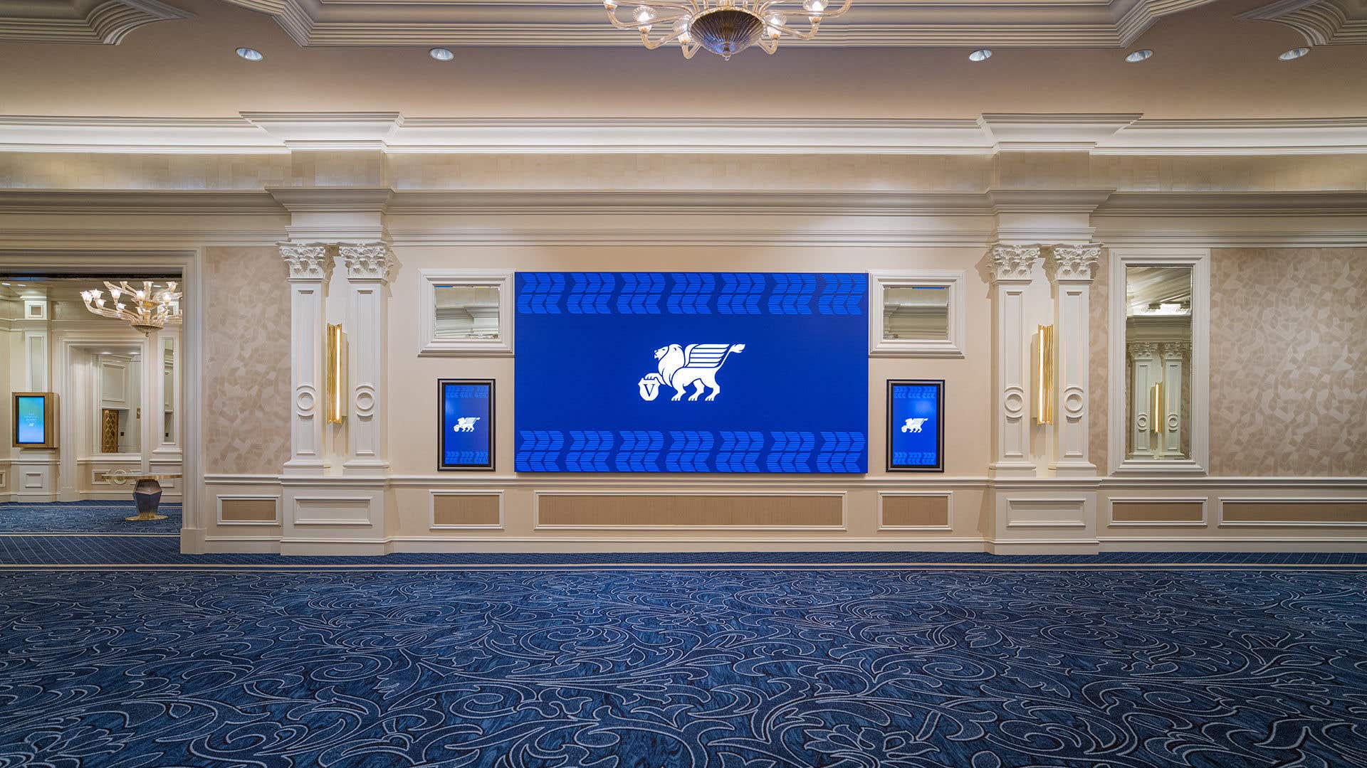 Elegant hallway with a large digital screen featuring a lion logo, surrounded by ornate columns, chandeliers, and blue patterned carpet.