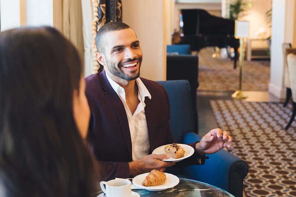 Two people sit at a cafe. One man smiles, holding a scone, while engaged in conversation.