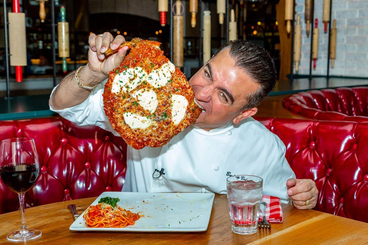 Person holding a large pizza  topped with melted cheese at a restaurant table.