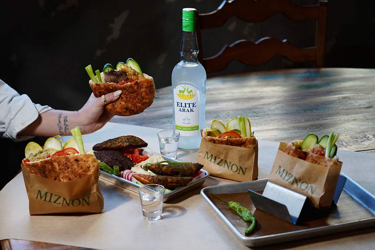An assortment of pita sandwiches on a table accompanied with a bottle of arak in the middle, with an arm on the left side of the picture holding up another pita sandwich
