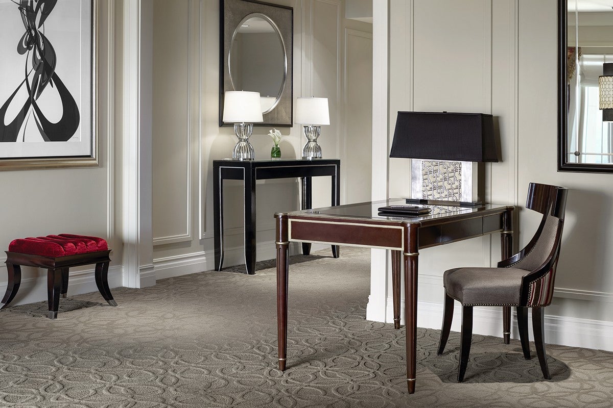Elegant hotel room with a writing desk, chair, decorative lamps, a mirror, and a bench, showcasing modern interior design.