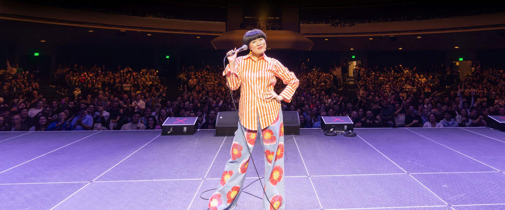 Atsuko Okatsuka in orange stripe top and floral pattern pants on stage holding a microphone with audience in the background.  