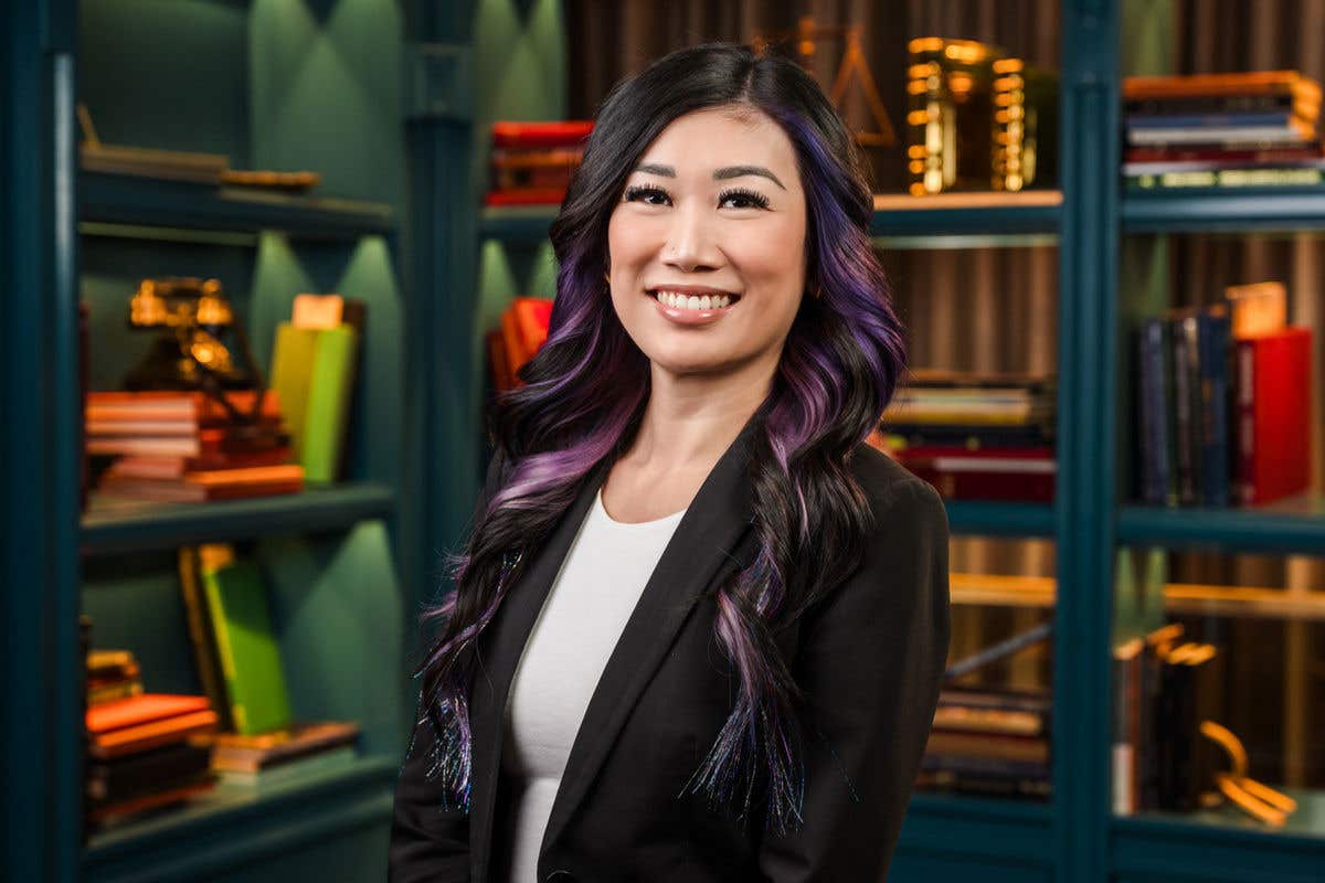 A professional headshot of a smiling woman with long, dark hair featuring purple highlights. She is wearing a black blazer over a white top, set against a teal bookshelf background.