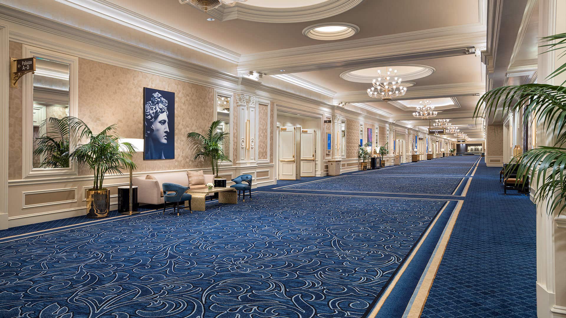 Elegant hallway with ornate chandeliers, blue patterned carpet, decorative wall panels, seating areas, and potted plants along the corridor.