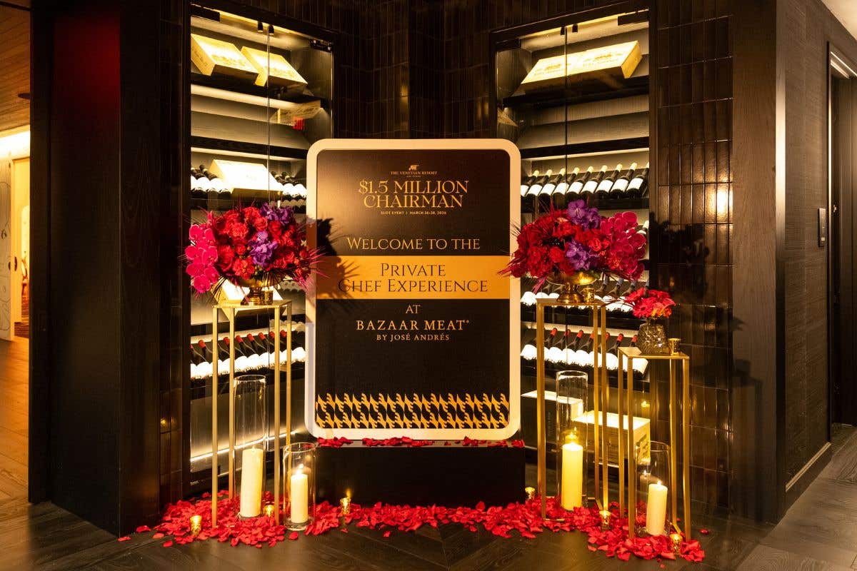 Elegant wine room display featuring a central sign for a "$1.5 Million Chairman Private Chef Experience at Bazaar Meat by José Andrés," surrounded by wine racks, red floral arrangements, candles, and rose petals.