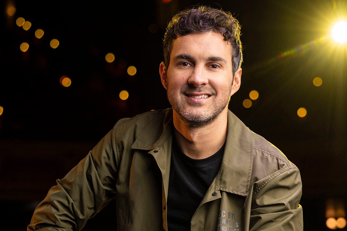 Mark Normand in a black shirt and olive/brown colored jacket against a black background with some out of focus lights and a bright yellow spotlight