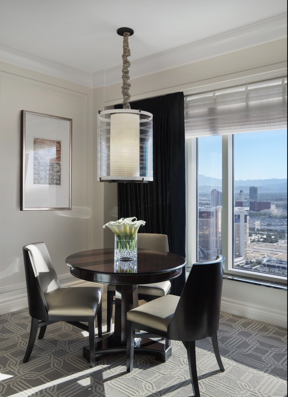 Round table with four chairs, a vase of white flowers, and a pendant light by a window with a city view in the background.