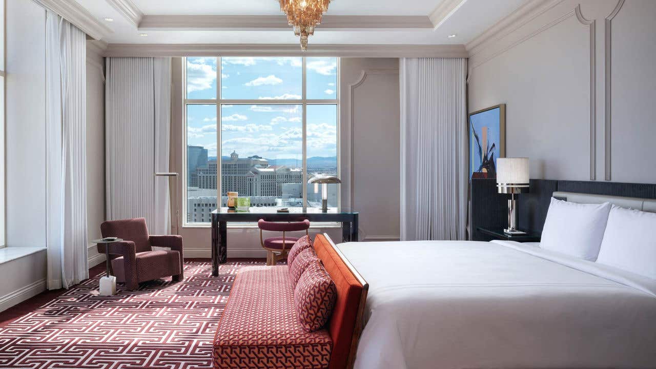 A corner hotel bedroom with a king bed, chandelier, desk, chair, bench, armchair, and two large windows with a view of the Las Vegas Strip.
