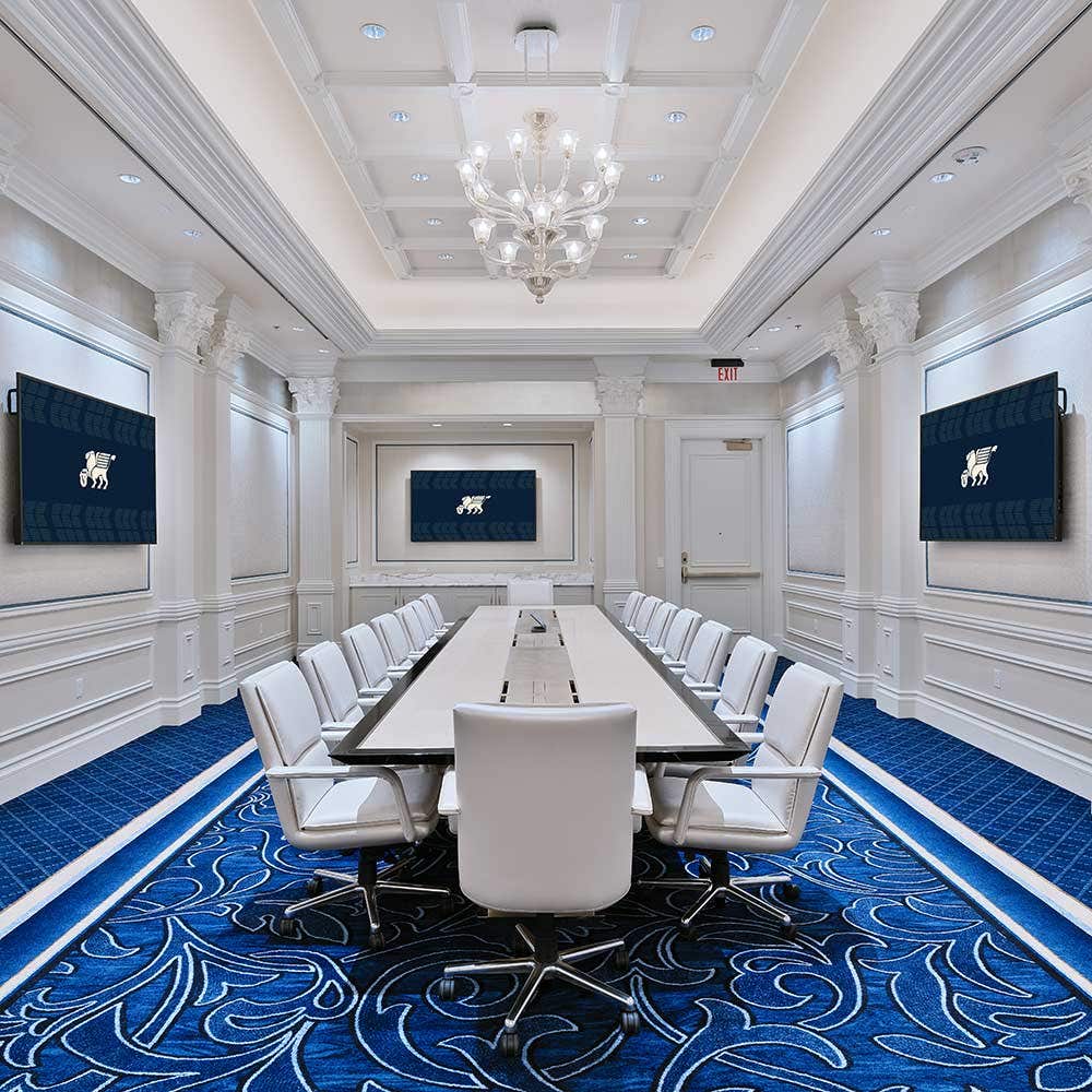 A table surrounded by chairs staged for a meeting set-up in a boardroom with a chandelier, blue carpet and three TVs hung on white walls.
