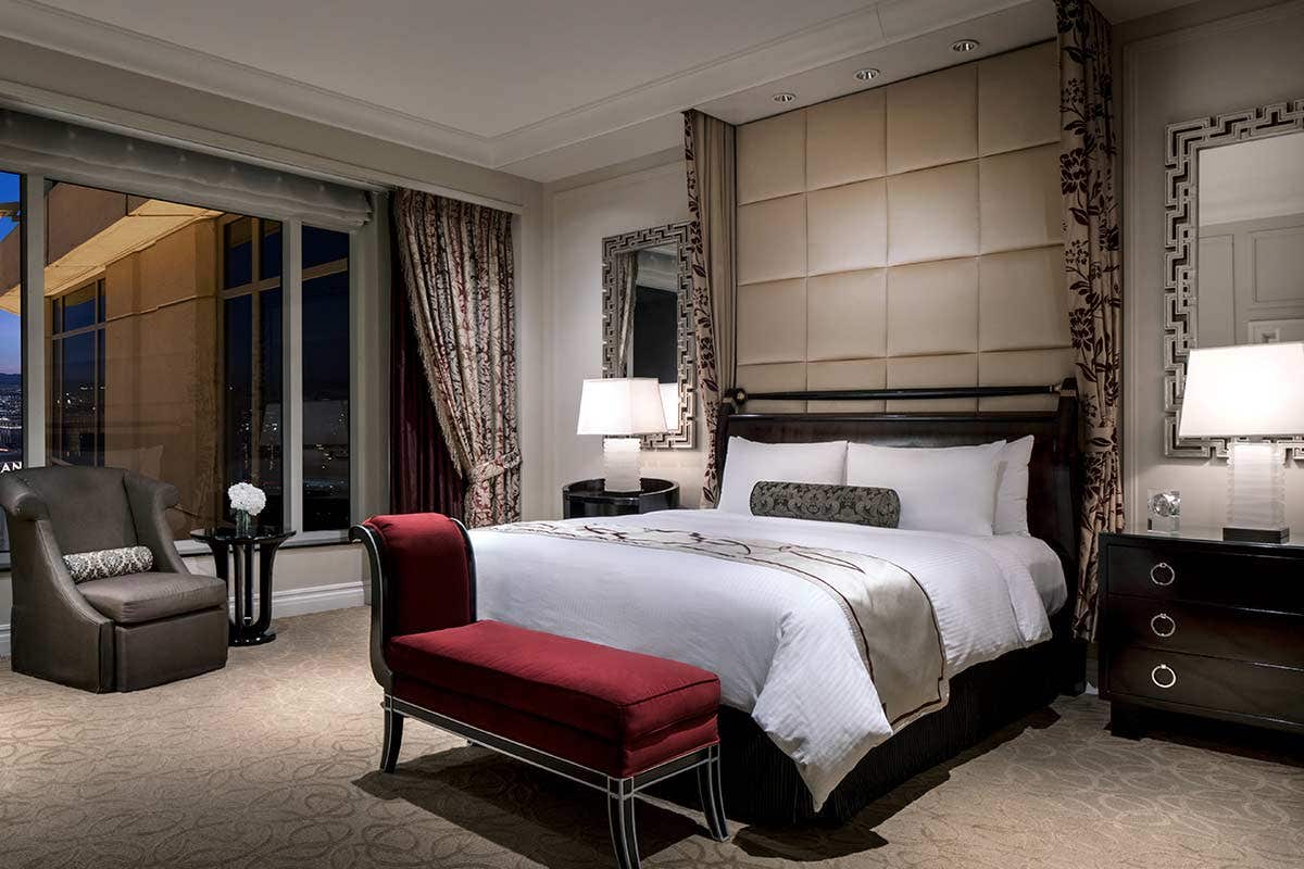 Luxurious hotel room with a king-sized bed, upholstered headboard, armchair, bedside tables, lamps, and city view window.