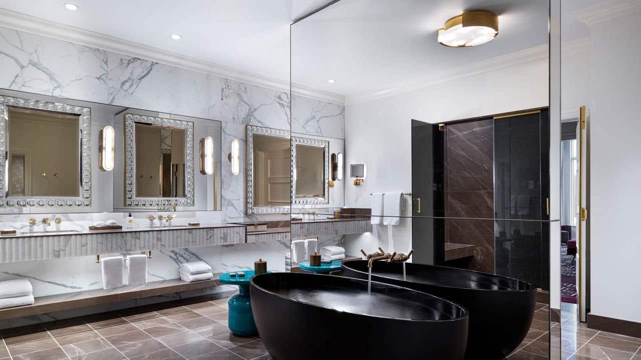 A hotel bathroom with a black freestanding bathtub, a mirrored separator wall, and a double sink vanity.