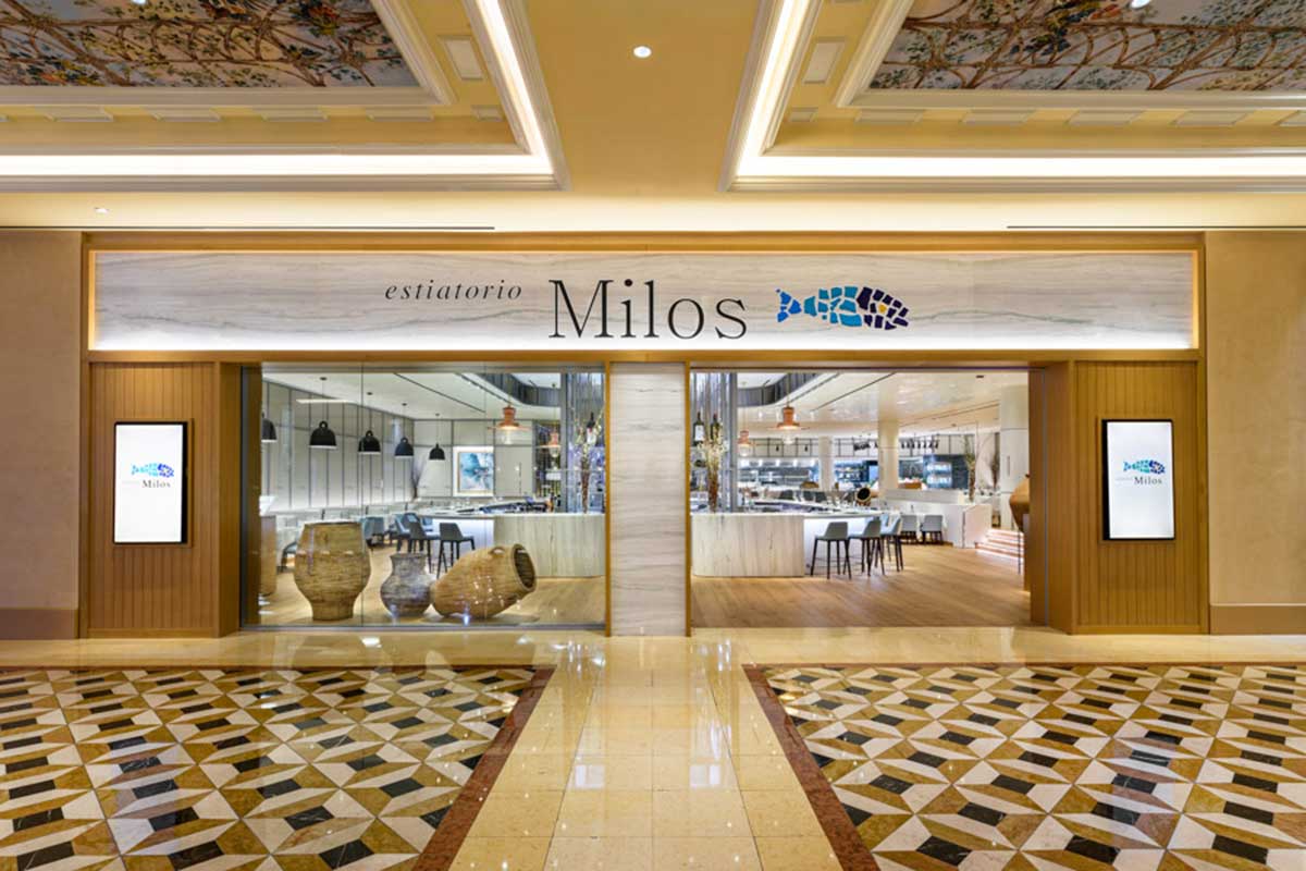 Entrance of Estiatorio Milos restaurant with modern decor, showcasing logo and interior seating through large glass doors.