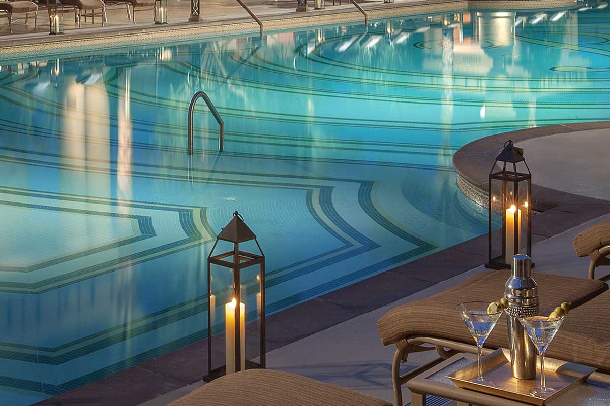 Close-up view of a resort pool at night with geometric tile patterns, showing two lounge chairs beside a small table holding two martini glasses and a shaker, with tall lanterns placed along the pool edge.