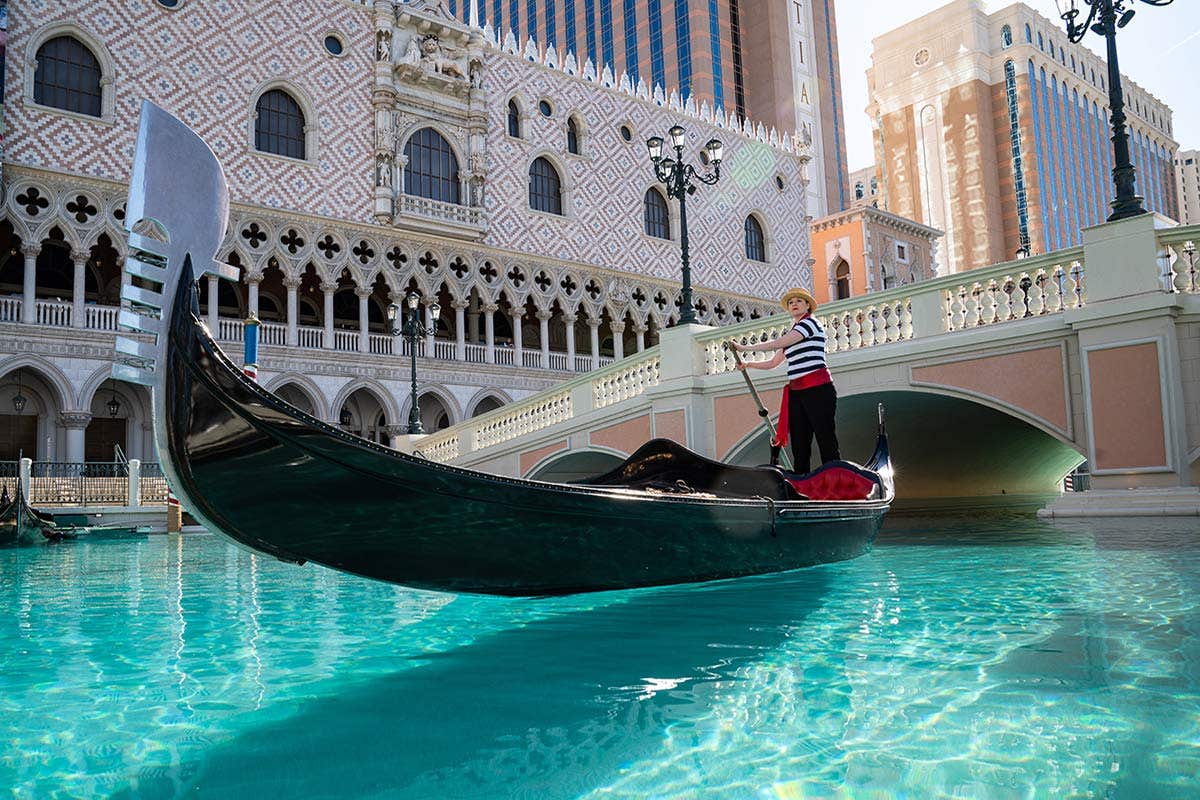 A gondolier navigates a sleek black gondola through a turquoise canal past ornate architecture and stone bridges at The Venetian Resort Las Vegas.