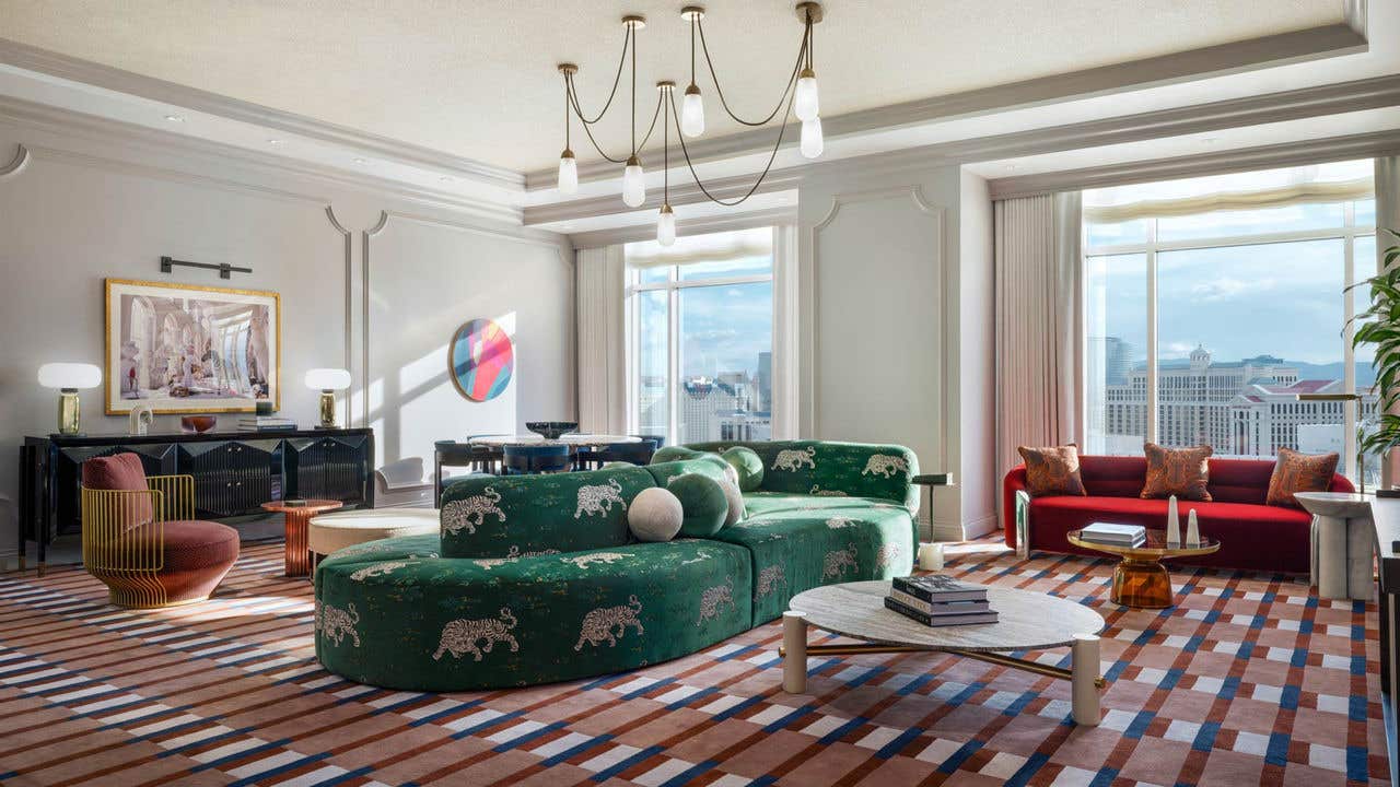 A hotel living room during the day with a double-sided dark green couch, chandelier, coffee table, love seat, dining table, armchair, and two large windows with a view of the Las Vegas Strip.