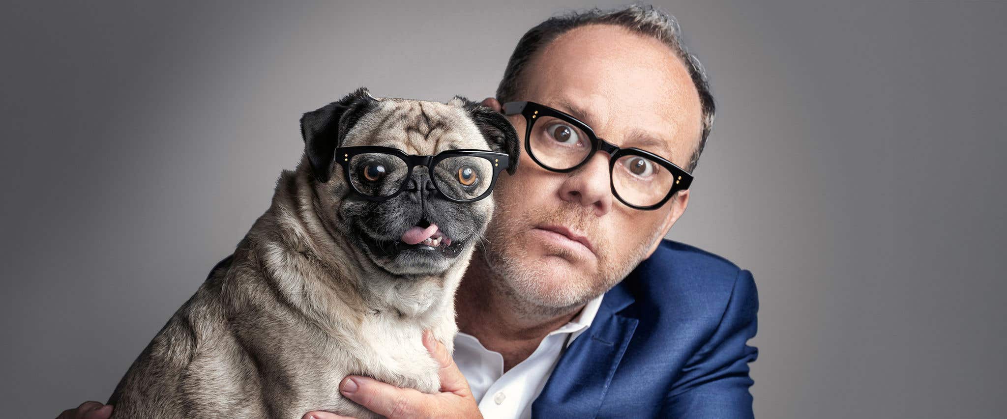 Tom Papa in blue jacket and white shirt holding a pug wearing glasses that look like his. 