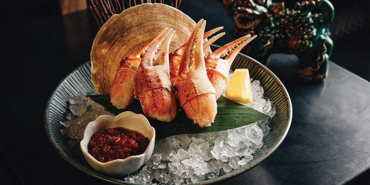 A group of crab claws over ice in a bowl with a side of red sauce and a lemon wedge on a restaurant table.