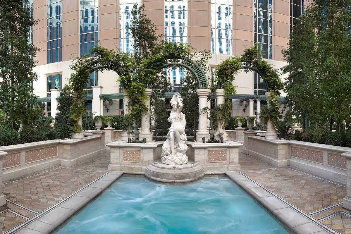 Courtyard featuring a carved stone statue surrounded by arched trellises covered in greenery, with a small turquoise pool in the foreground and a tall hotel building in the background.