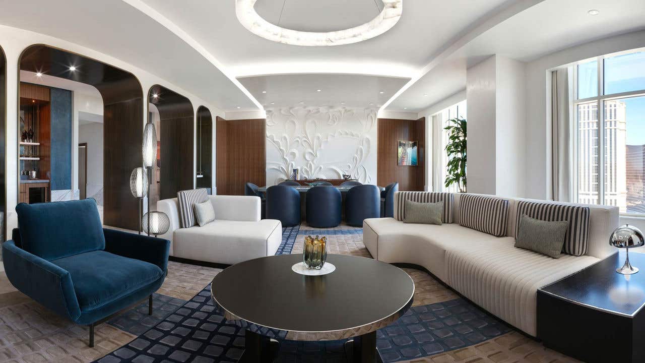 A white and blue hotel living room with a couch, coffee table, chairs, and a dining table with eight chairs.