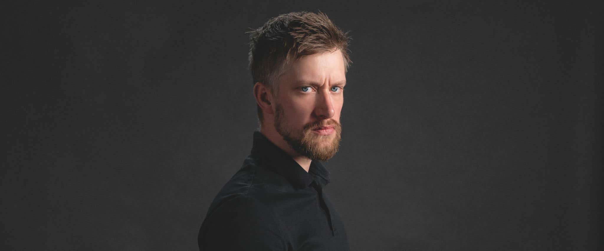 Daniel Sloss in a black shirt standing sideways in front of a black background staring directly at the camera. 