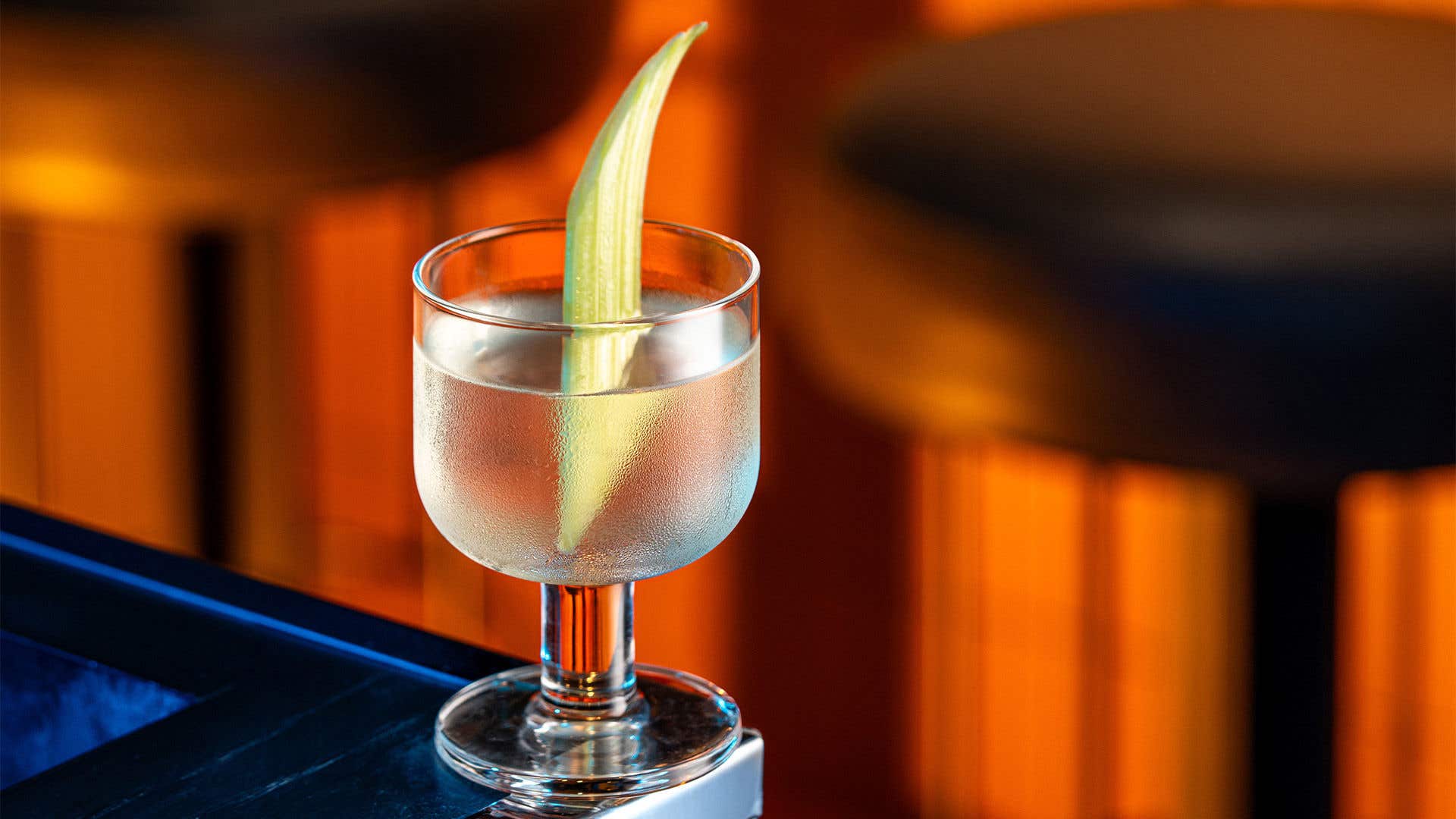 Cocktail with clear liquor in a small glass on a table with a celery garnish. 