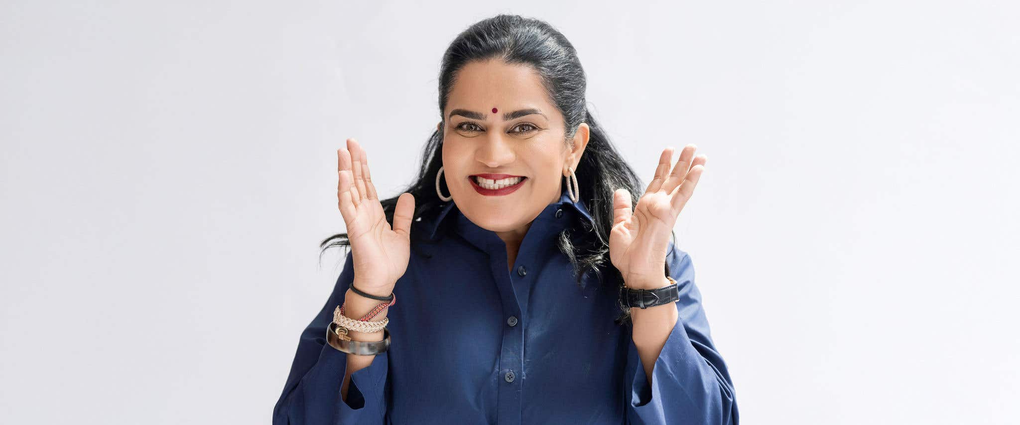 Zarna Garg in a blue dress standing in front of a white background smiling at the camera with her hands raised up to her face. 