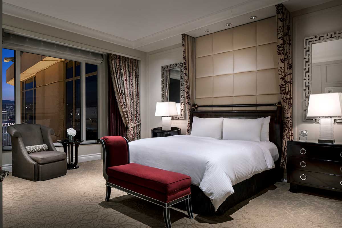 Hotel room at night with a king-sized bed with white linens and a plush headboard, flanked by nightstands with lamps and a red bench at the foot of the bed. A window with a city view on the left with an armchair and side table in front of it.  