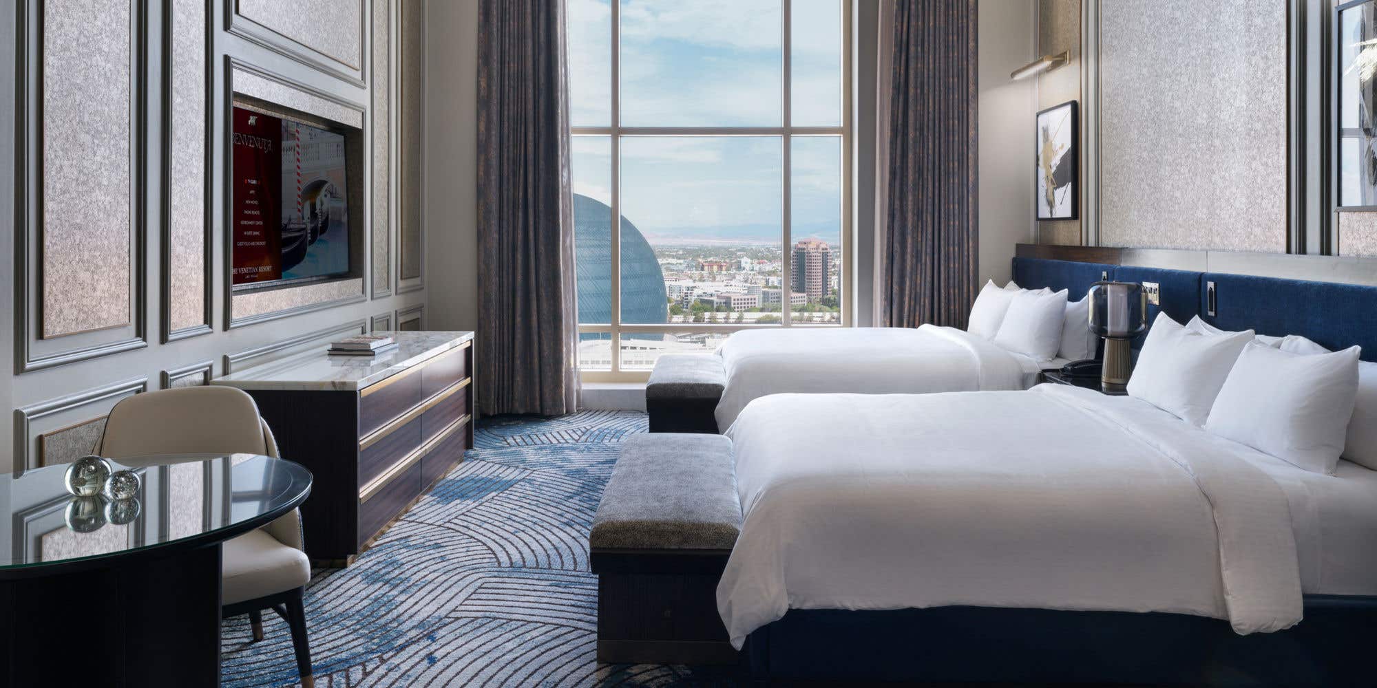 A hotel room with two queen beds, a table, a TV, a chandelier, and a tall arched window with a view of the Sphere.