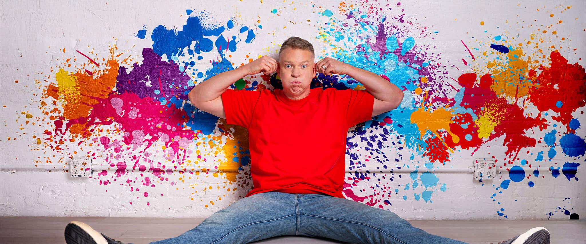 Gary Owen in a red shirt and jeans sitting against a white wall with paint splatters on it pulling at his ears. 