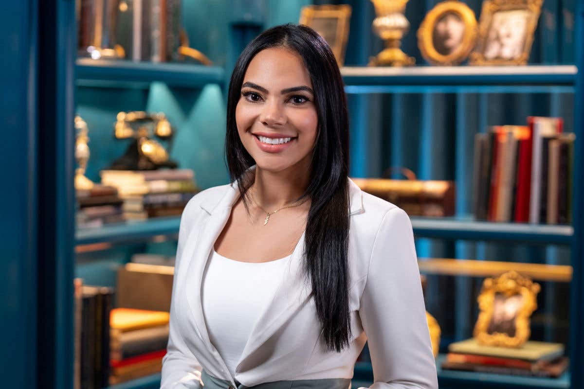 A professional headshot of a smiling woman with long dark hair, wearing a white blazer. She is standing in front of a blue bookshelf filled with books and vintage decor.