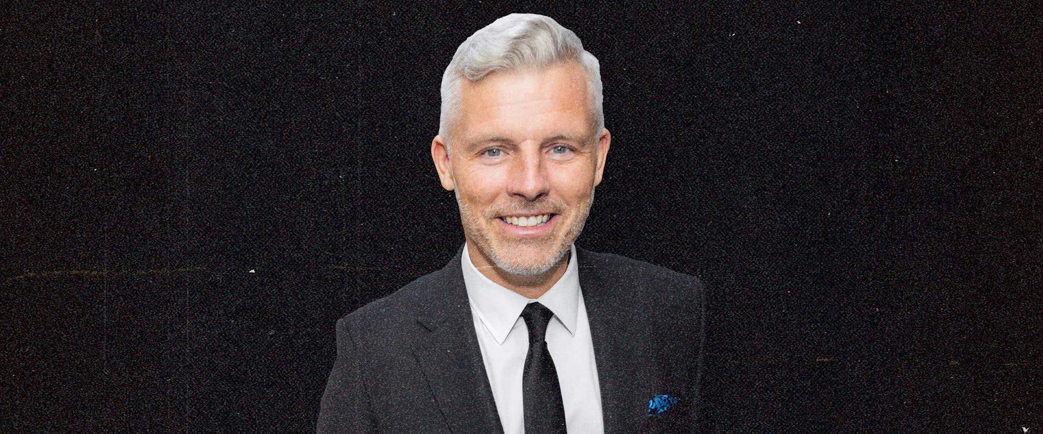 Des Bishop in front of a black background wearing a black suit with black tie smiling at the camera.