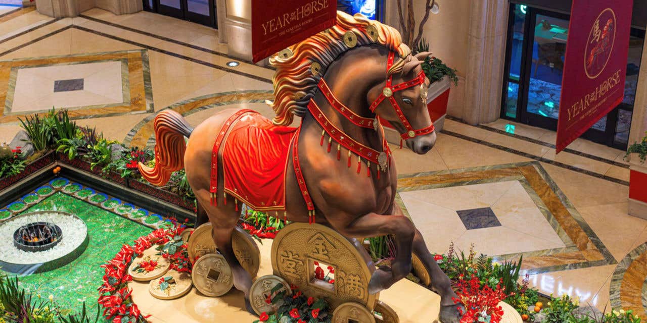 An ornate brown horse statue stands at the center of an indoor display, decorated with a red and gold saddle and stepping onto giant gold Chinese coins. Surrounded by lush plants and red flowers, the scene is framed by banners marking the Year of the Horse.