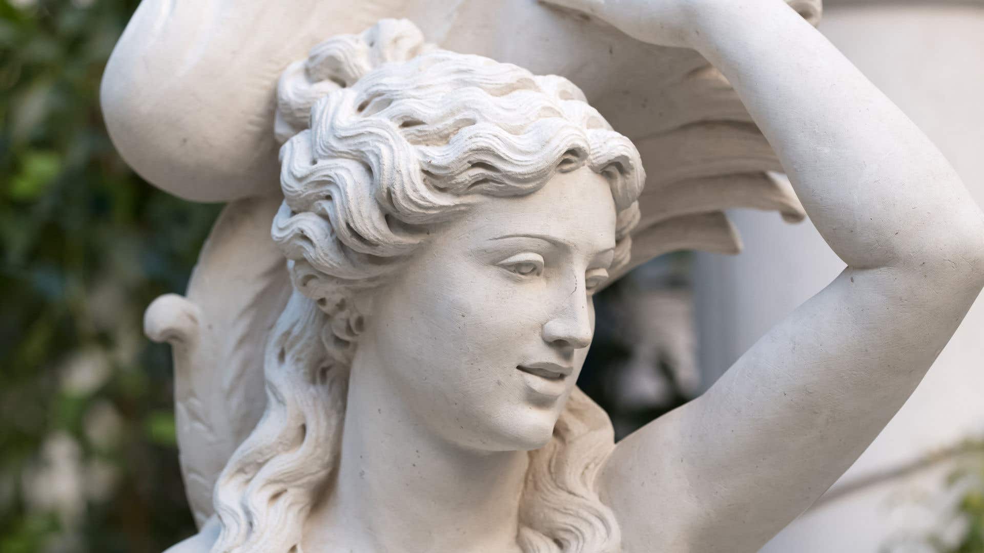 Close-up view of a carved stone statue featuring detailed wavy hair and an arm raised overhead, with soft natural greenery visible in the background.