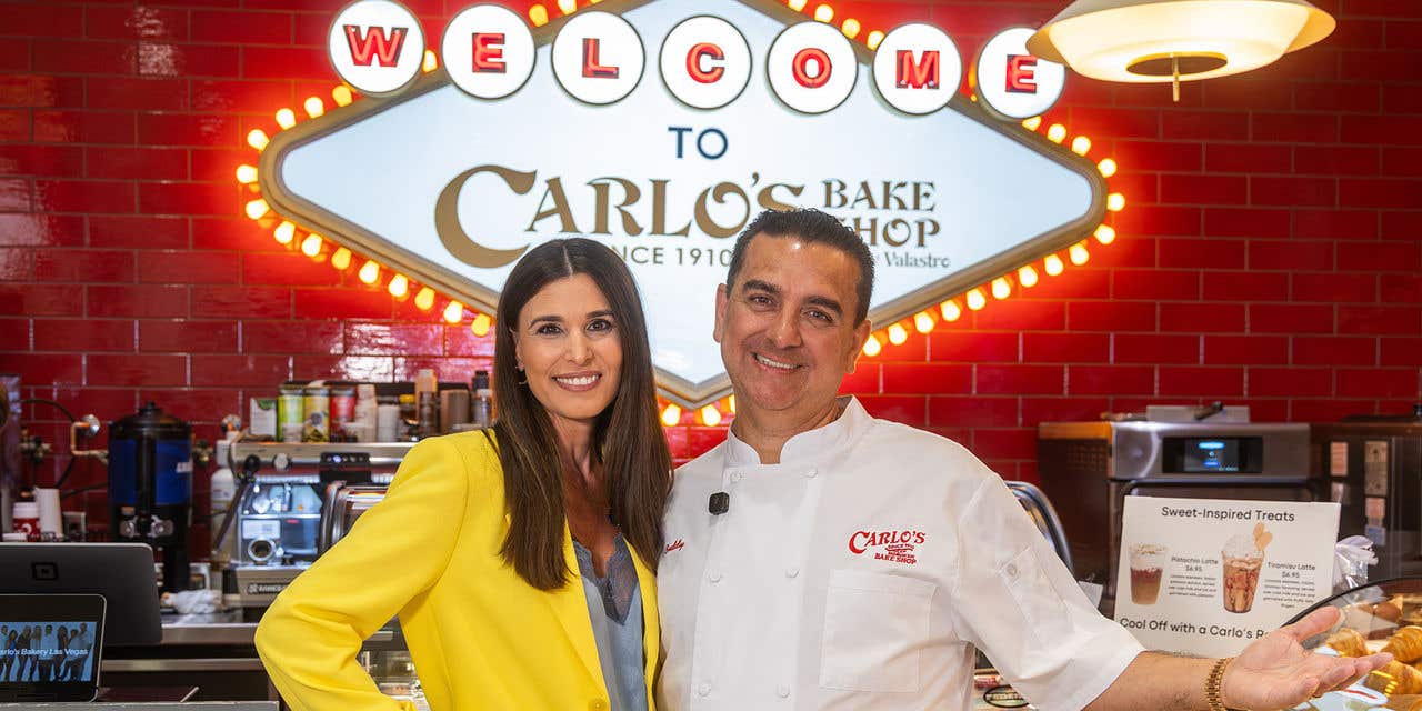 Two people pose inside Carlo’s Bake Shop beneath a bright welcome sign.