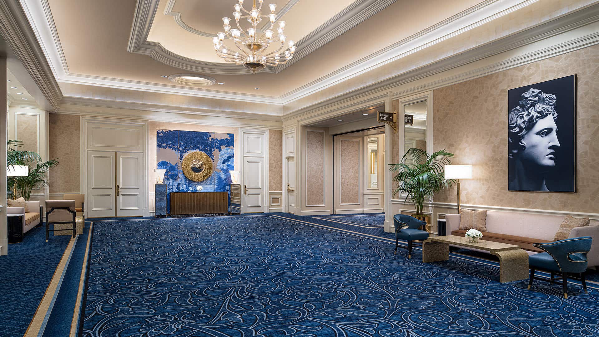 Elegant hallway with blue patterned carpet, chandeliers, ornate walls, a seating area with sofas, and decorative artwork and plants.