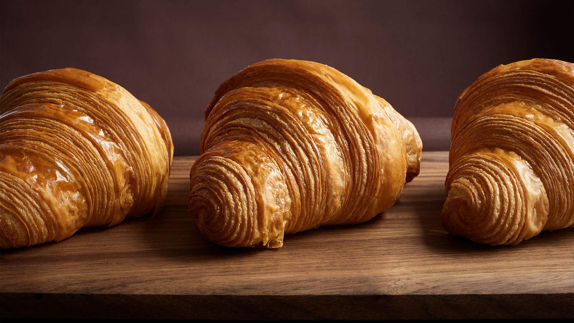 Three golden, flaky croissants arranged in a row on a dark wooden board.